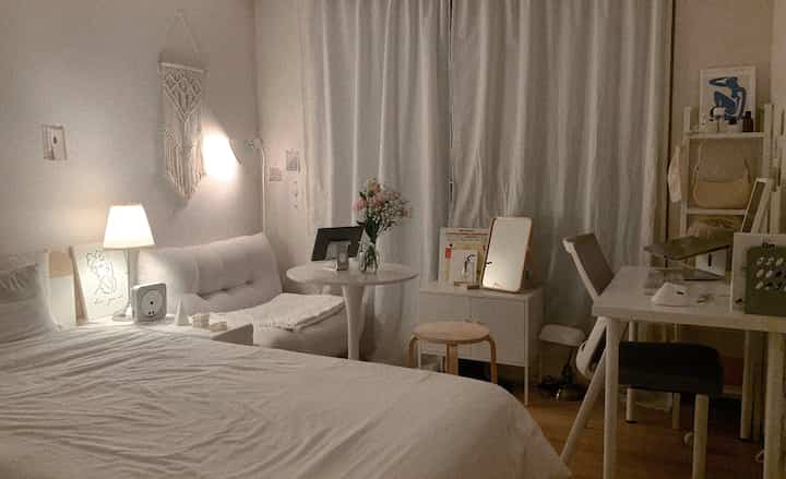 A cozy small bedroom in white and beige tones featuring a bed, sofa, and desk with warm lighting