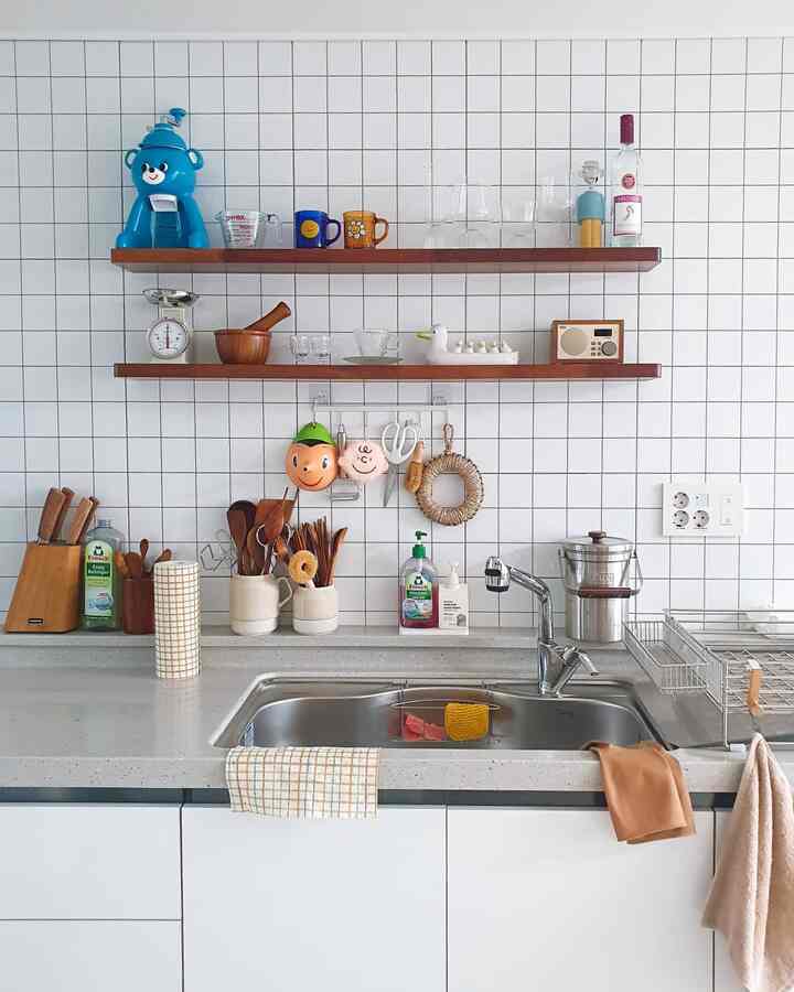 白いタイルの壁と木製の棚があるナチュラルなキッチン、シンク周りにキッチンタオルと調理器具が整った清潔感のある空間