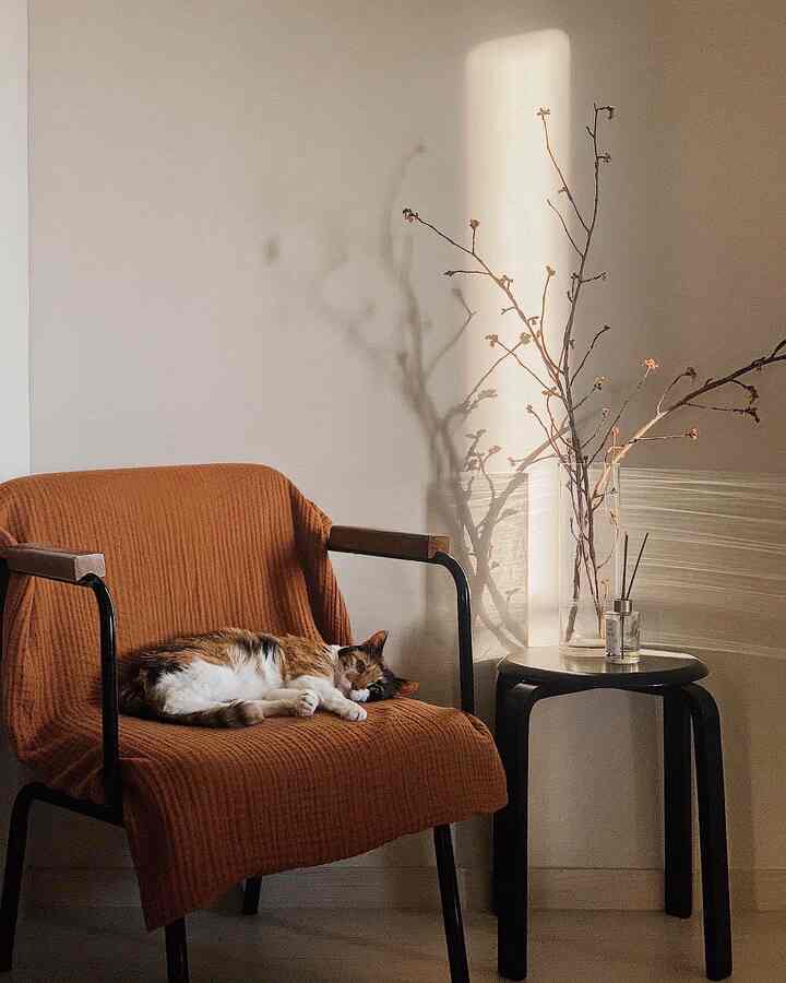 Natural-toned bedroom reading nook featuring a brown blanket-covered armchair and a black stool with diffuser and vase creating a cozy atmosphere