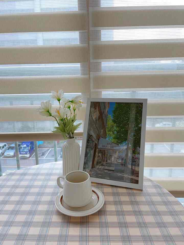 ベージュ調のバルコニー空間、チェック柄のテーブルクロスの上に白いカップと花瓶、額縁が置かれた雰囲気のあるおうちカフェ