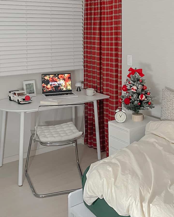 White and red toned bedroom with home office setup featuring a transparent chair and a small decorated Christmas tree in a neat interior