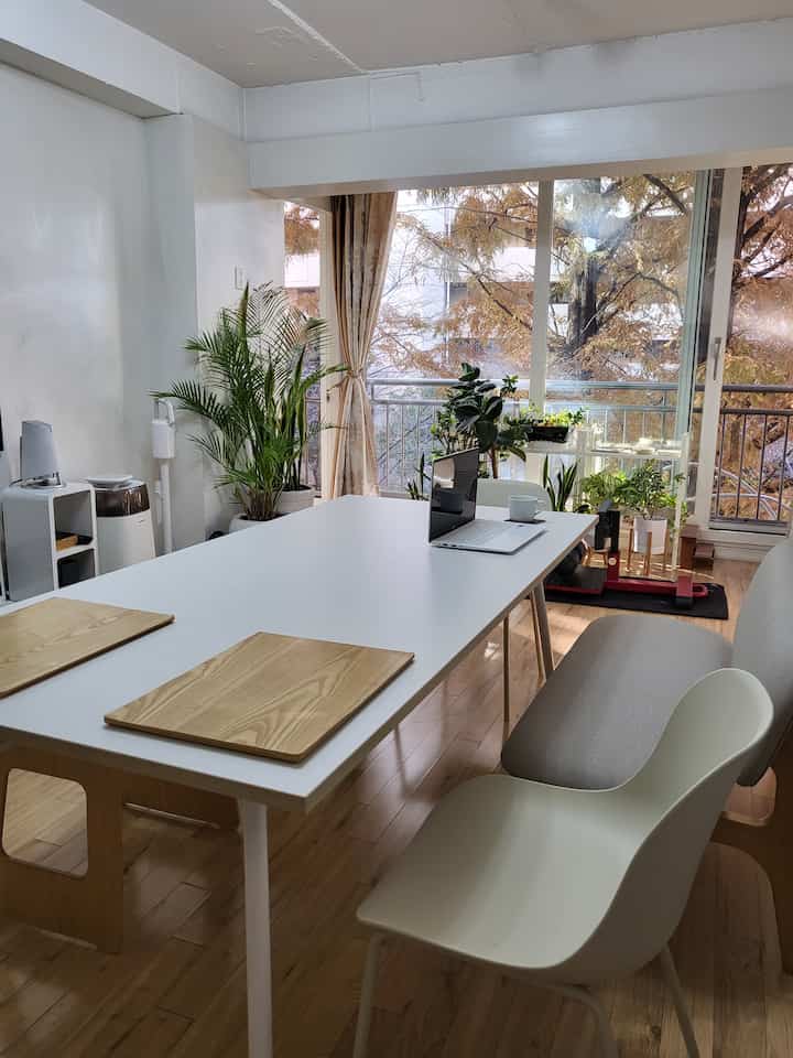 Natural wood and white toned living room style home office featuring a large dining table, chairs, bench, and abundant plants, creating a cozy, natural atmosphere