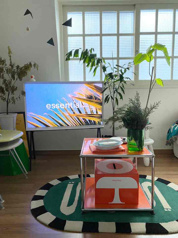 White-walled living room with wood tone floor, featuring an orange coffee table, TV stand, and multiple plants creating a simple natural ambiance