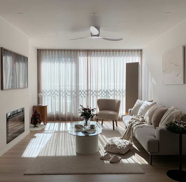 A natural beige-toned living room featuring large windows with beige curtains, an armchair, and sofa creating a cozy and simple atmosphere
