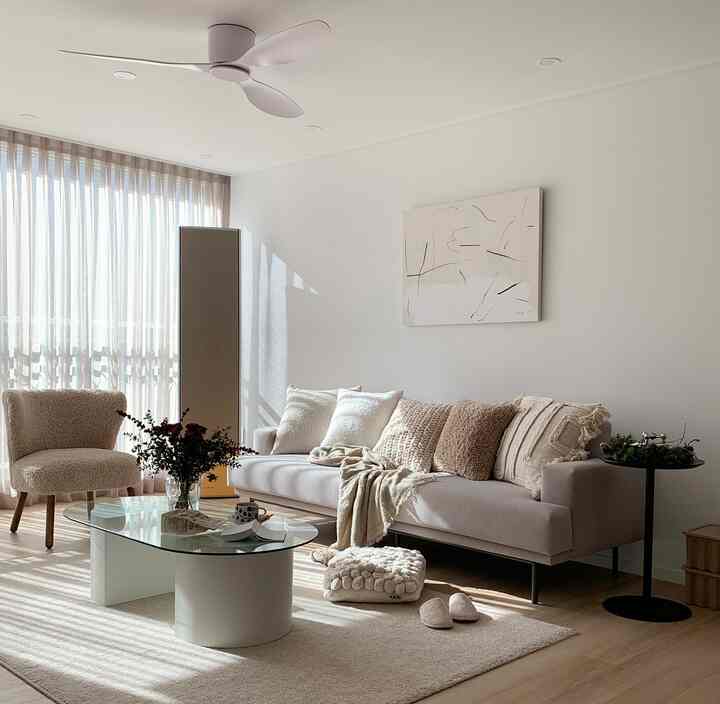 Bright beige and white toned living room featuring beige sofa and armchair with glass-top coffee table in warm ambiance