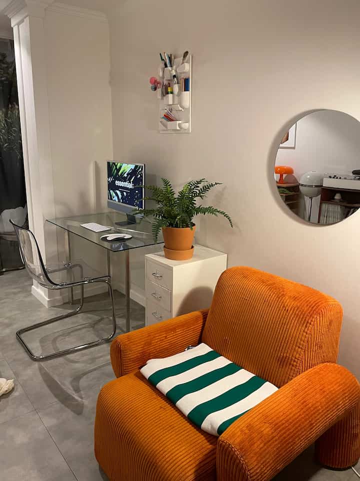 A cozy home office space with white walls and gray tiled floor, featuring a transparent chair, orange armchair, and potted plant on a glass desk.
