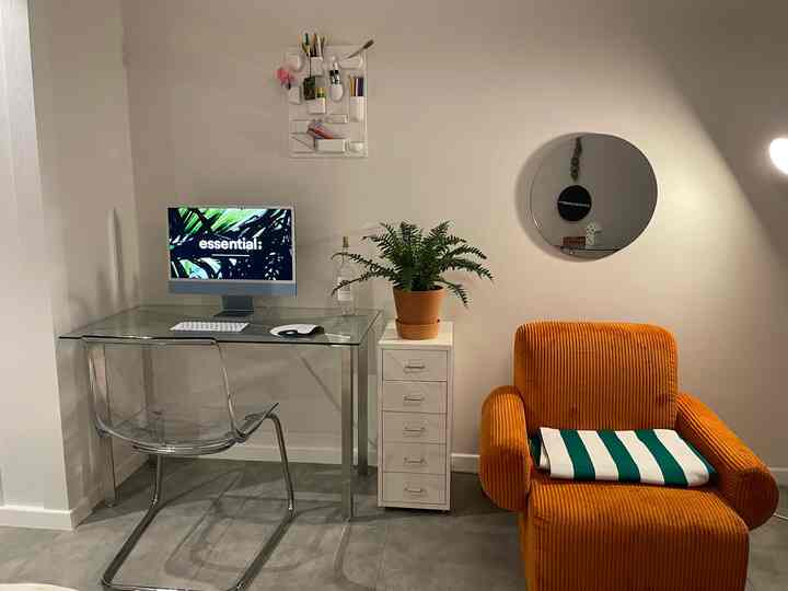 Modern home office with white walls and transparent chair, featuring an orange armchair and plant creating a cozy atmosphere