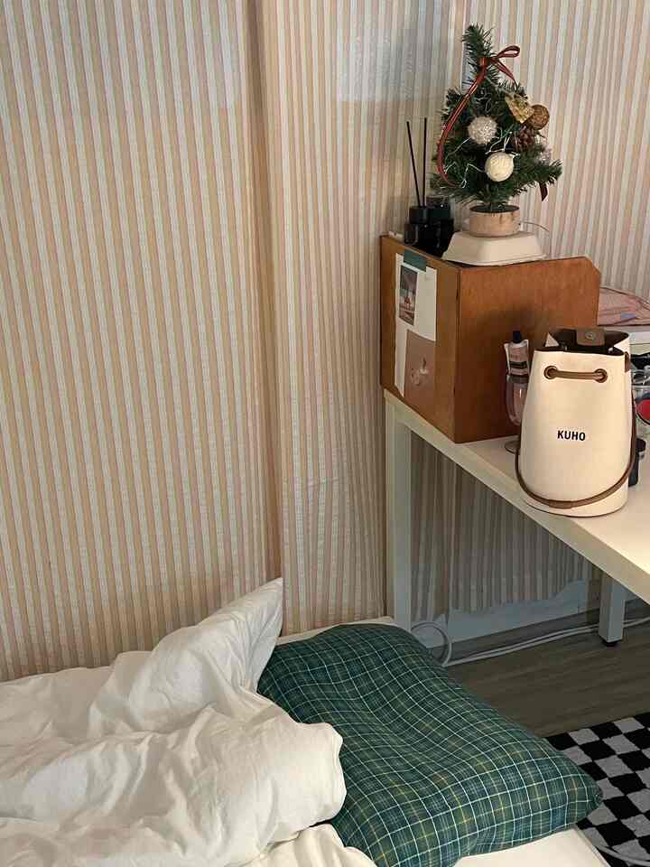 白と木目調の調和がある寝室、窓際にベッドとチェック柄の枕、小さな机の上にミニクリスマスツリーが置かれた雰囲気のあるインテリア