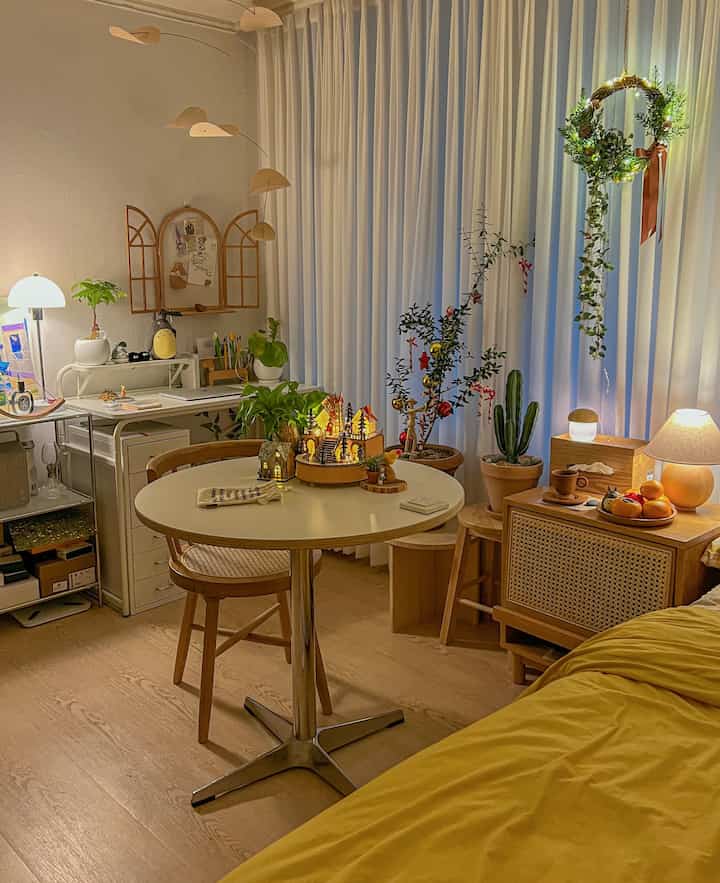 Natural wood tones and white curtains in a studio apartment, featuring a dining table, nightstand, and many plants creating a warm atmosphere