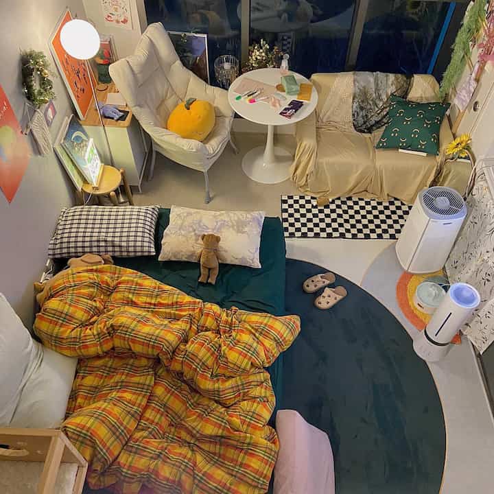 A small bedroom with white walls and soft lighting, featuring yellow plaid bedding, round rugs, an armchair, and posters creating a cozy atmosphere