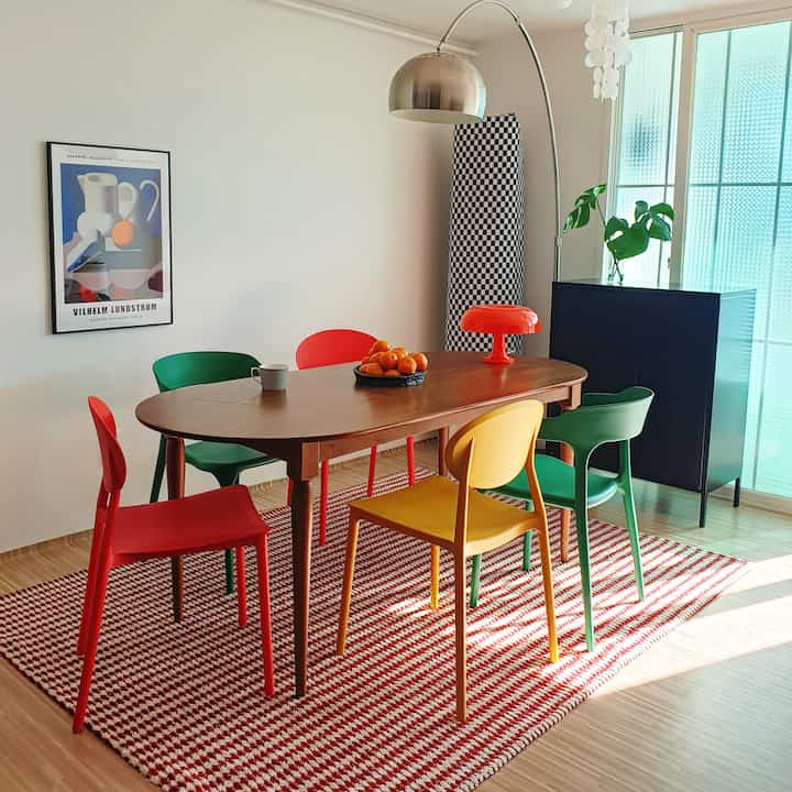 Wood tone dining room featuring red and green dining chairs with a mid-century modern poster and floor lamp, creating a simple atmosphere