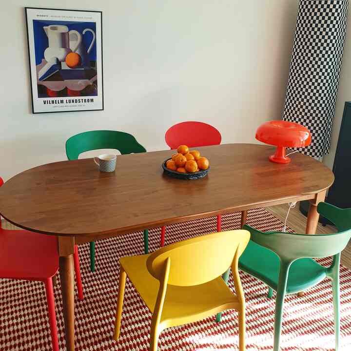 Cozy dining room featuring a wooden table surrounded by red, green, and yellow chairs, bright lighting, and a modern poster creating a warm atmosphere