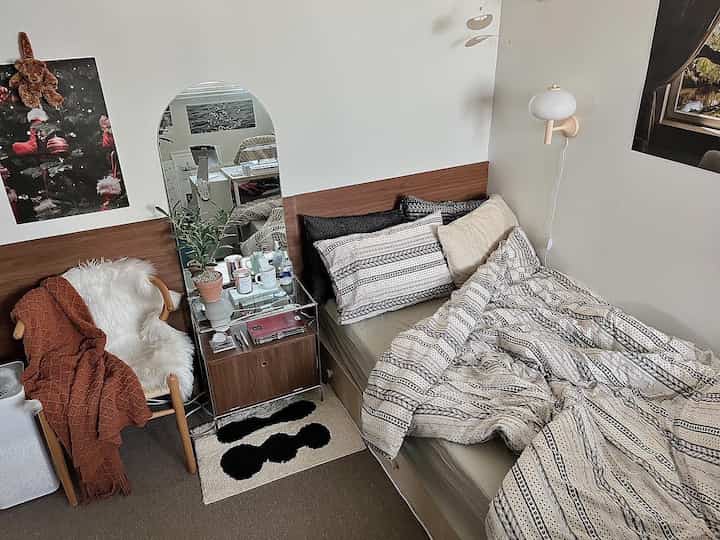 Natural-toned 6-tatami bedroom featuring a bed, nightstand with potted plant, arched mirror, and wooden armchair creating a cozy space