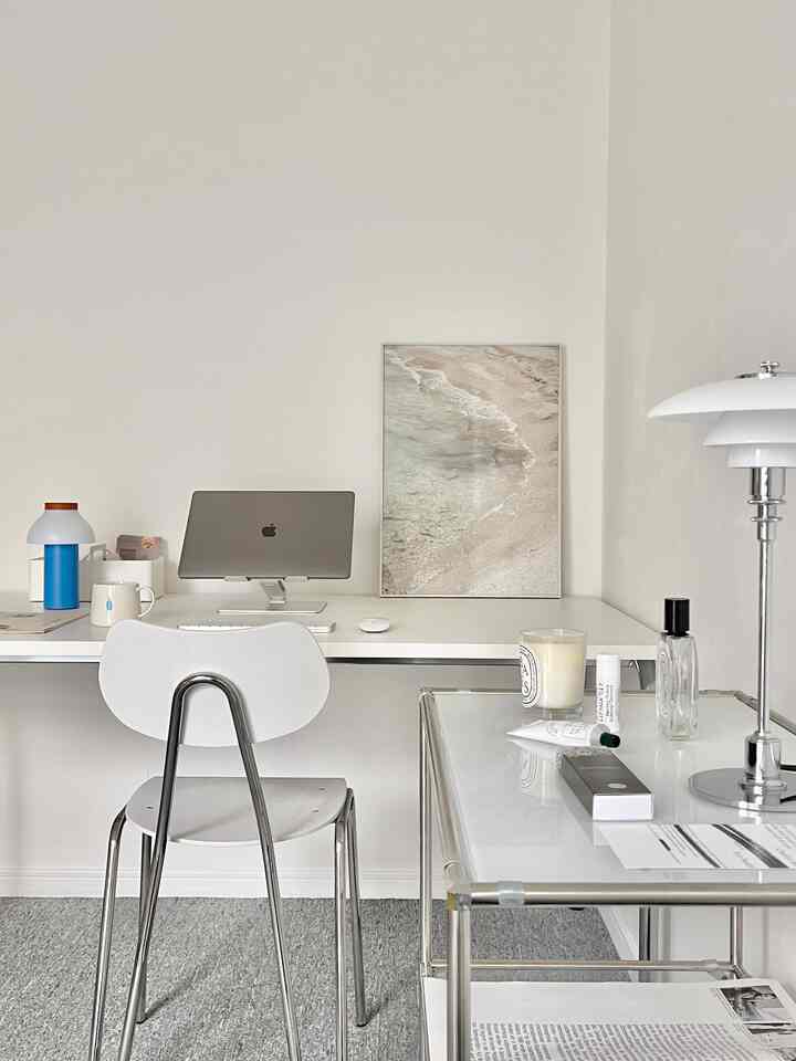 A compact home office space featuring white and silver-toned furniture, gray carpet, and a beach poster creating a clean working atmosphere