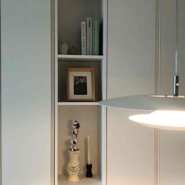 Bright and calm white-toned dining room featuring shelving next to table, wooden picture frame, cotton vase, and pendant lighting creating warm atmosphere