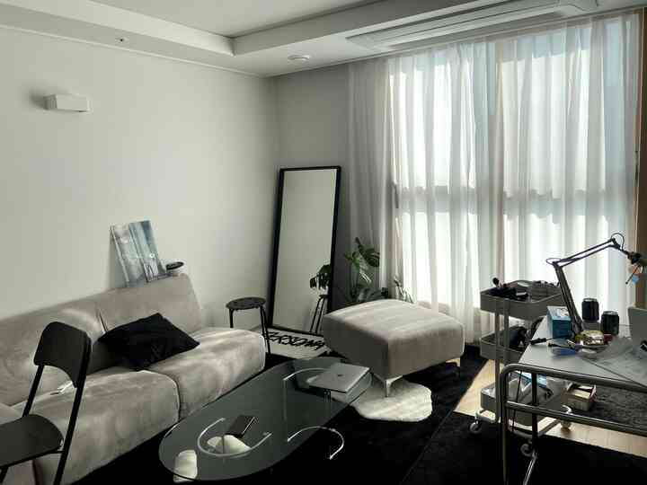 明るいグレーとブラックのトーンが調和した窓辺のリビング、ソファとオットマン、大きなシフォンカーテンがあるモダンでコンパクトな一人暮らし空間