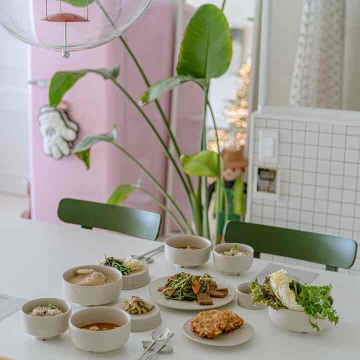 柔らかいホワイトとグリーンの色調のダイニング、中央にダイニングテーブルと椅子、多様な家庭料理と大きな観葉植物が配された雰囲気のある空間
