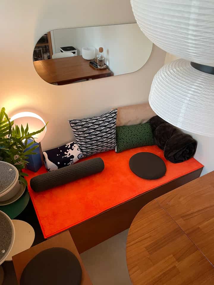 Warm-toned living room featuring a wood tone bench with bright orange cushions and assorted patterned pillows, a long wall mirror, and a potted plant