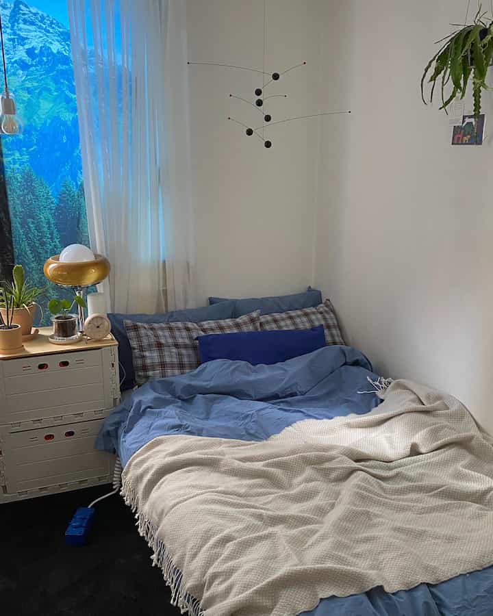 Blue-toned studio bedroom featuring a bed with pillows and blankets centered, with plants and lighting on the left side creating a cozy atmosphere