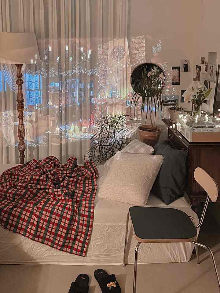 Warm beige-toned studio bedroom featuring a mattress with a checkered throw, plants, and a projector casting imagery on sheer curtains, creating a cozy home cinema vibe