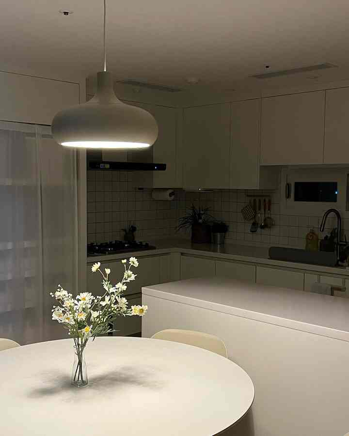 White-toned kitchen space featuring a round dining table, pendant light, and artificial flowers creating a clean and simple atmosphere