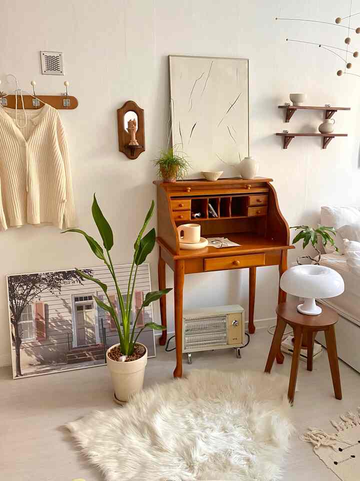 Cozy studio apartment with warm wood tones, featuring a vintage roll-top desk, stool, ivory rug, and indoor plants.