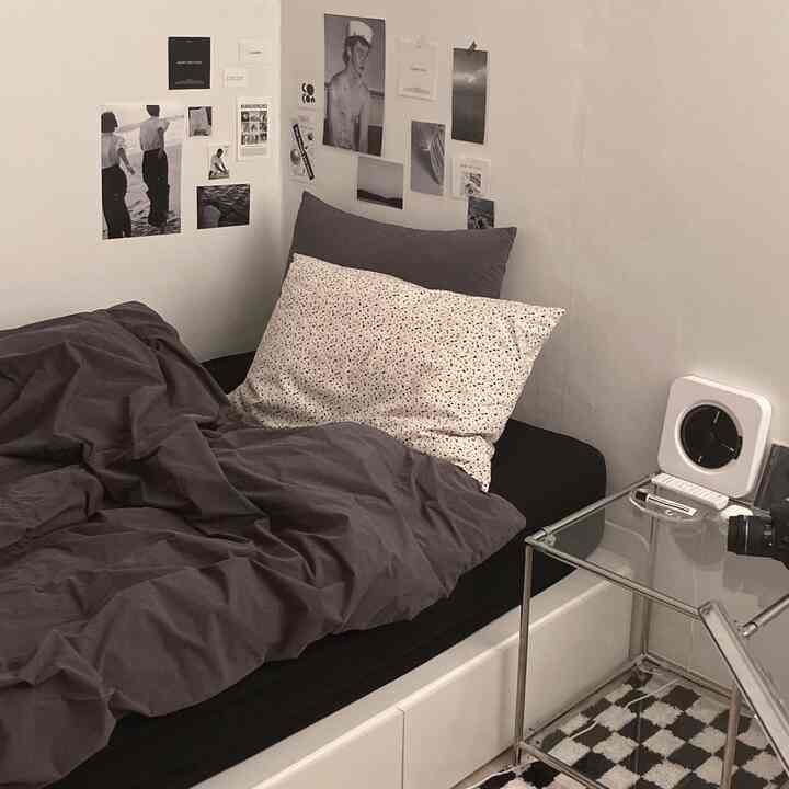 モノトーンの寝室空間、ダークグレーの寝具とチェック柄のラグ、透明なサイドテーブルと多数のポスターが配置された落ち着いたモダンなインテリア