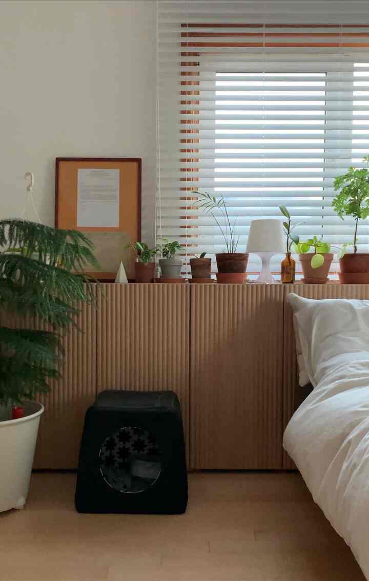 木目調のブラインドとナイトテーブル上に観葉植物が配置された白い寝室のインテリア