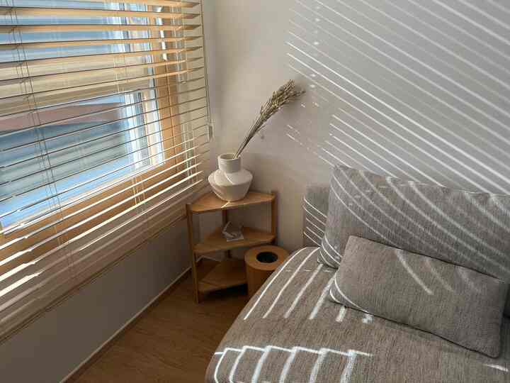 ナチュラルな木目調のブラインドと木製コーナーシェルフ、ベージュのソファが配置された居心地の良いリビングの隅の空間