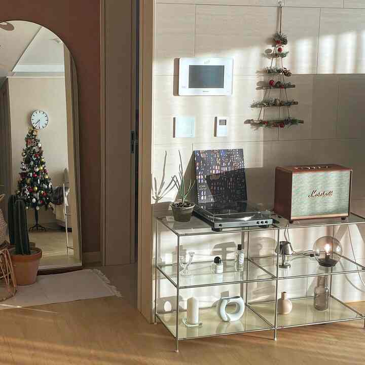 暖かいブラウンの木製床とホワイトタイルの壁があるリビングコーナー、ガラス収納棚の上にターンテーブルとスピーカーがあり、左側の鏡にクリスマスツリーが映っている印象的な空間