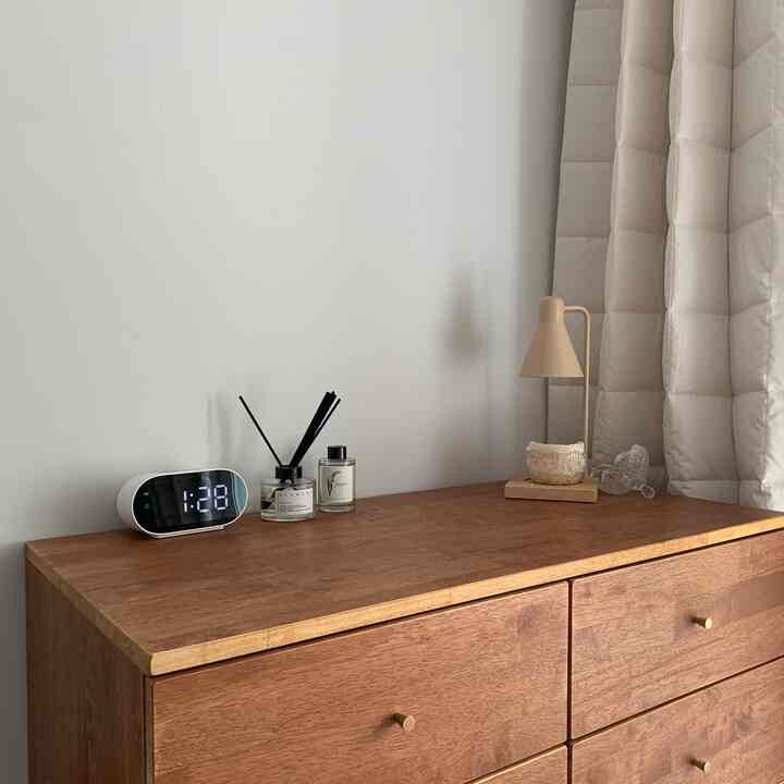 Warm and simple space featuring a brown wooden dresser with digital clock, diffuser, and beige-toned lamp on top