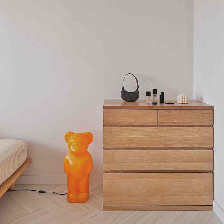 Natural wood tone bedroom featuring a dresser and bed with a clean minimal design, highlighted by an orange bear-shaped lamp and cosmetics on the dresser