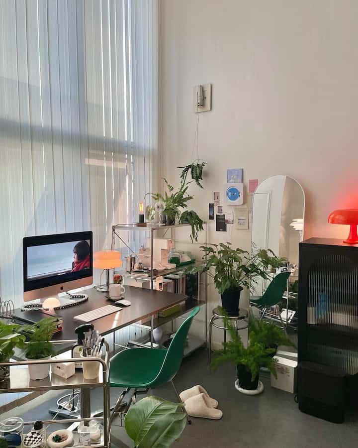 白い壁とカーテンを背景にしたコンパクトなホームオフィス、黒い机と緑の椅子、多数の植物が配されたミニマルでモダンなインテリア