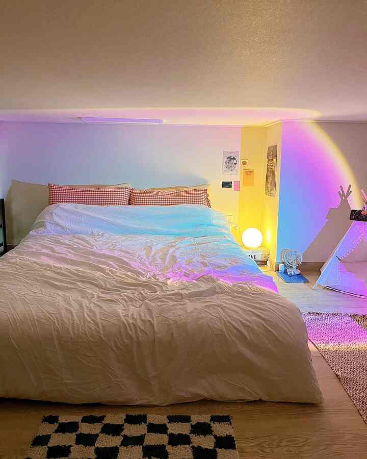 A cozy studio bedroom with warm yellow and purple lighting, checkered pillows on bed, a round mood lamp, and a dog pet tent arranged warmly
