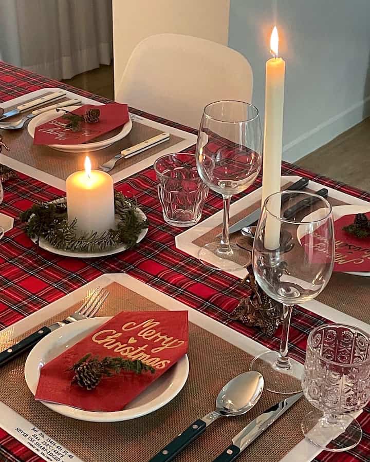 A dining room table with red plaid tablecloth and Christmas decorations, featuring wine glasses and lit candles creating a warm atmosphere.
