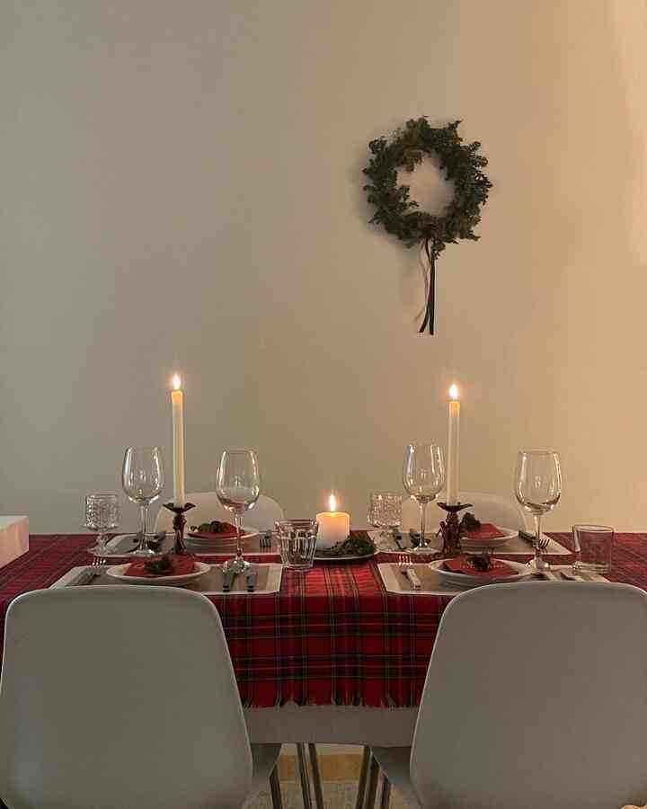 Dining room with red plaid tablecloth and white chairs, featuring a Christmas wreath and candles creating a cozy home party atmosphere