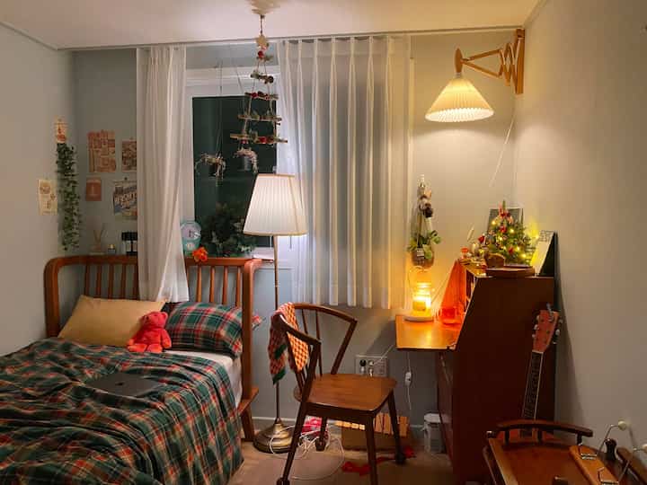 Cozy small bedroom with wood tones and red-green checkered bedding, featuring Christmas decorations and antique furniture.