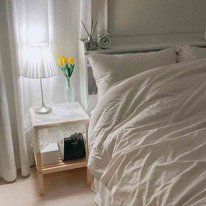 明るい白を基調とした寝室、木製ナイトテーブルにレースのテーブルクロスと黄色のチューリップの花瓶、テーブルランプが温かみのある雰囲気を演出するインテリア