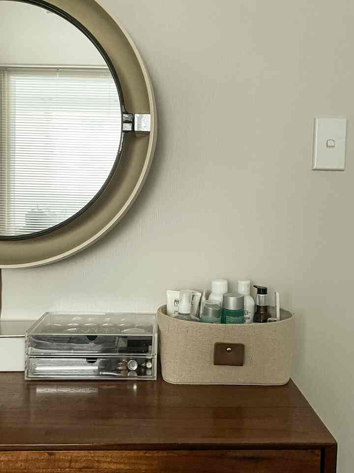Natural tone living space with round mirror and dresser featuring transparent organizer and beige fabric storage basket