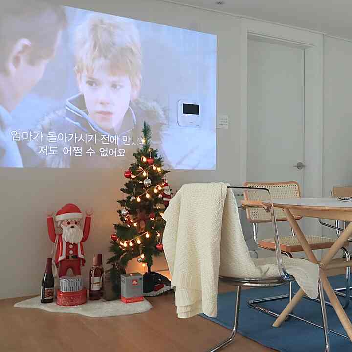 白い壁と木の床に青いラグが敷かれたダイニングルーム、ダイニングテーブルとラタンチェア3脚、クリスマスツリーとサンタのフィギュアが雰囲気のあるホームシネマ空間を演出