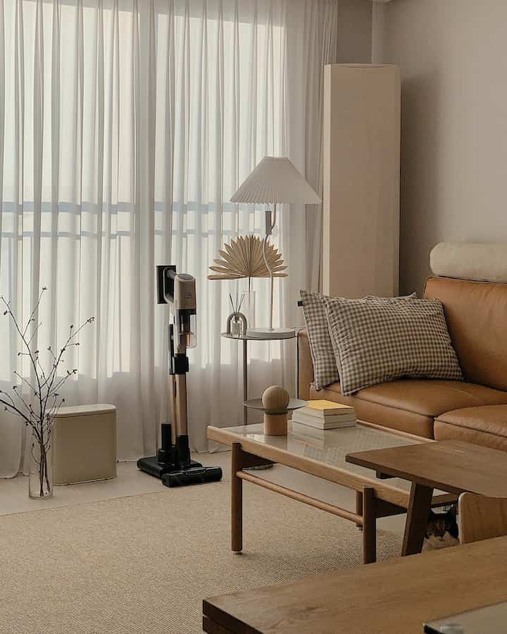 Natural wood tone and beige living room featuring leather sofa, wireless vacuum, and curtains in a cozy setting