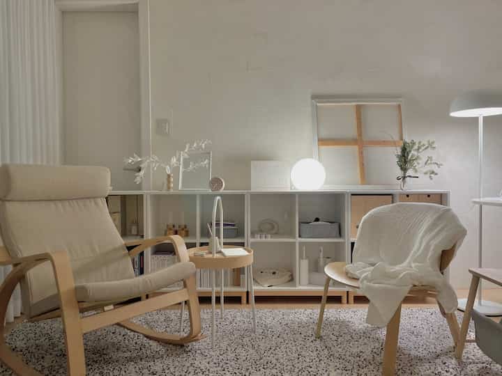 ホワイトと木目調が調和したワンルームのリビング、アームチェアと本棚が静かでミニマルな雰囲気の空間に配置されている