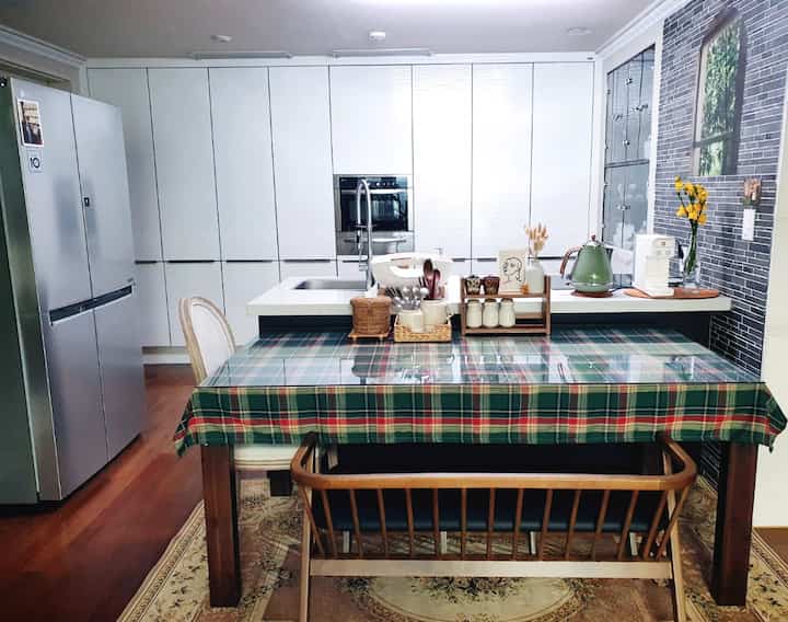 白い壁面収納と木目調のダイニングテーブルがあるナチュラルな雰囲気のキッチン、チェックのテーブルクロスと黄色いフリージアがアクセント