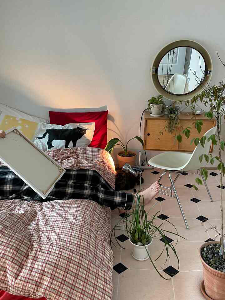 Beige floored bedroom with red bedding, featuring plants, a round mirror, and a white armchair creating a cozy atmosphere