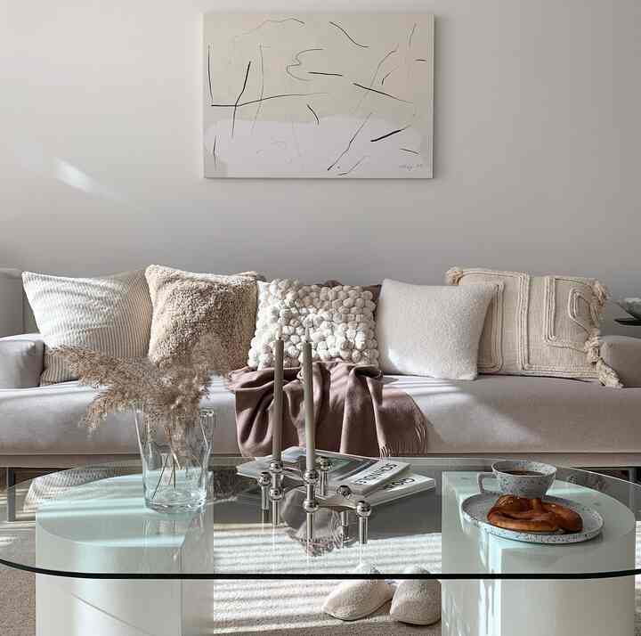 Bright white and beige toned living room featuring beige sofa, textured cushions, glass coffee table, and objets creating a natural atmosphere