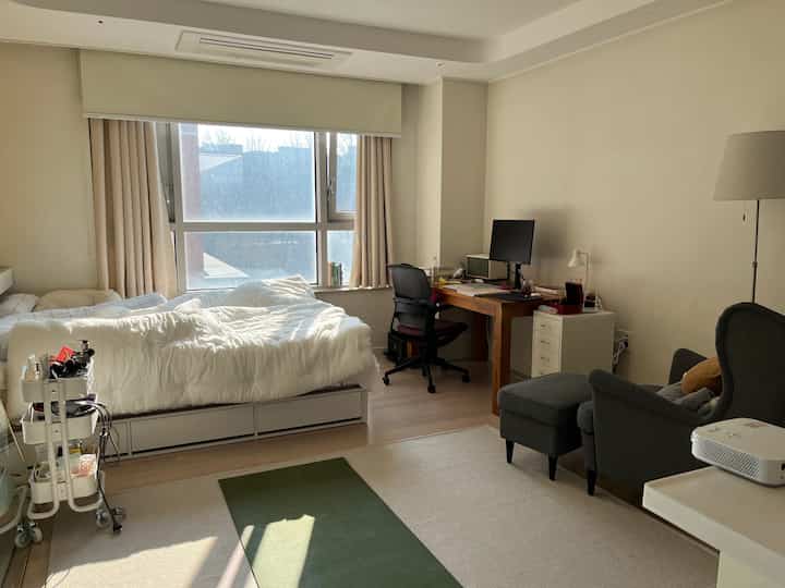 Beige and white toned single household bedroom and home office space featuring a bed by the window and wooden desk, creating a natural atmosphere
