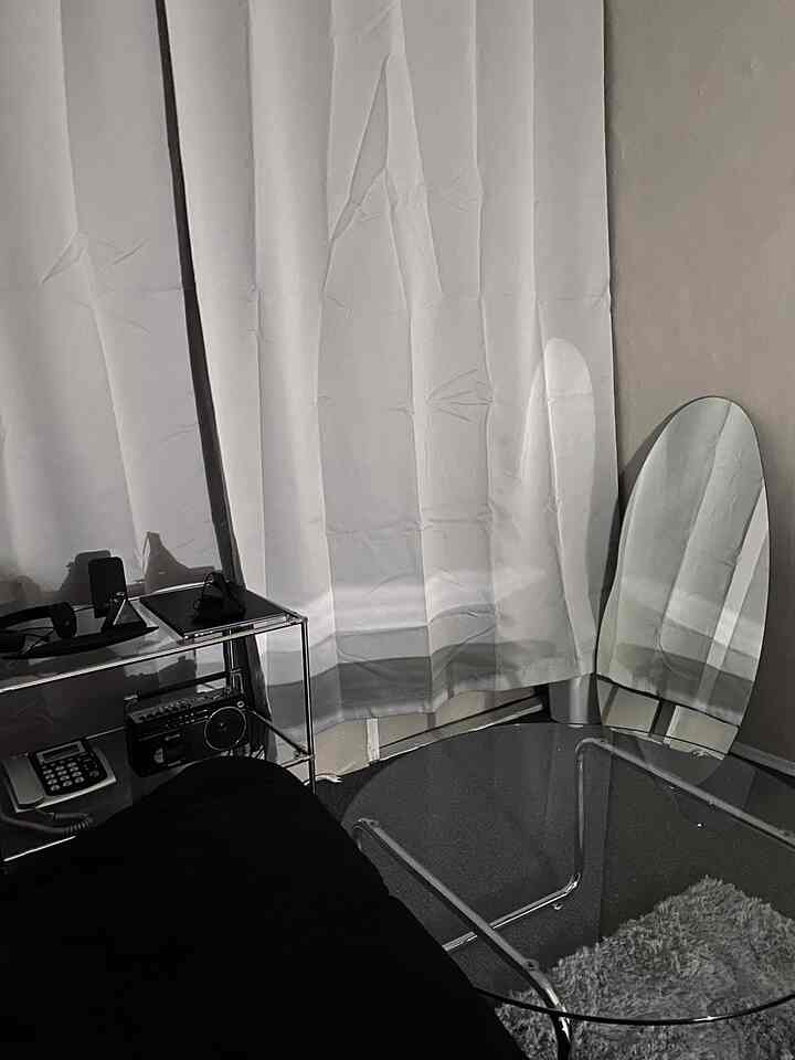 Small room interior featuring black sofa, metal frame furniture, glass table, and white curtains