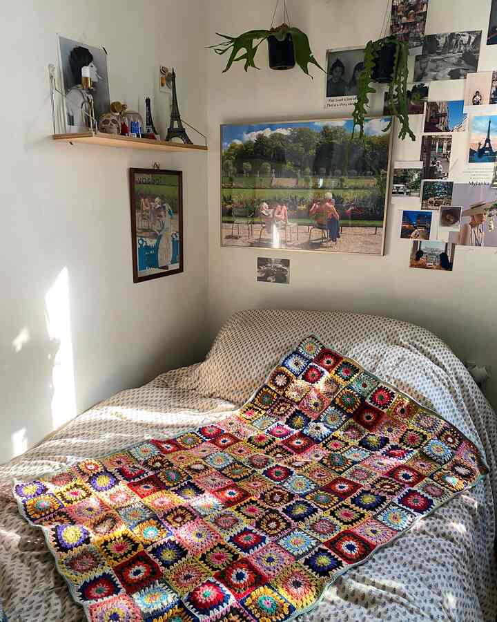 白いトーンの寝室にカラフルなヴィンテージのかぎ針編みスローがベッドに掛けられ、壁にはヴィンテージスタイルのポスターと写真が飾られた雰囲気のあるワンルームインテリア