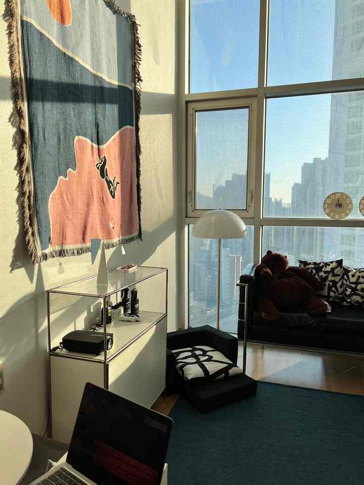 A modern loft living room bathed in warm natural light, featuring a black sofa with large teddy bear and a colorful wall tapestry