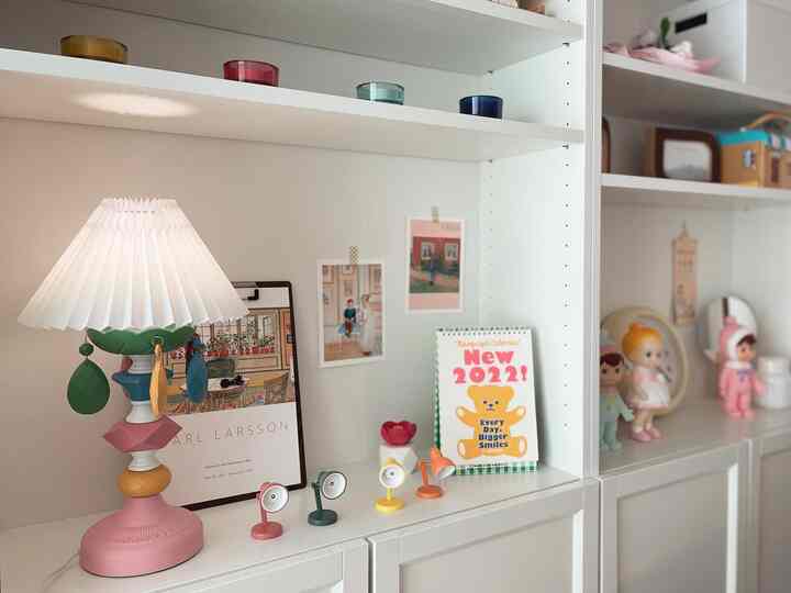 White-toned kids' room bookshelf featuring pastel-colored lamps, cute dolls, and colorful posters creating a cozy atmosphere